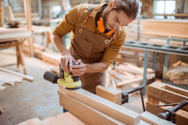 Carpenter grinding wooden bar with a cordless grinding machine in the workshop