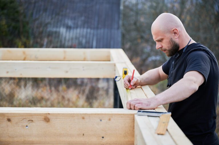 Man worker building wooden frame house on pile foundation. Bald carpenter using tape measure for measuring wooden planks and making marks with pencil. Carpentry concept.